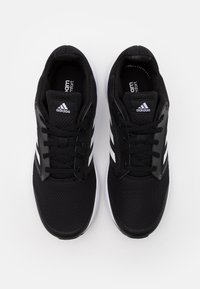 Svarta träningsskor med ovandel i mesh, som har tre vita ränder, en svart plast hälkappa och en vit sula.