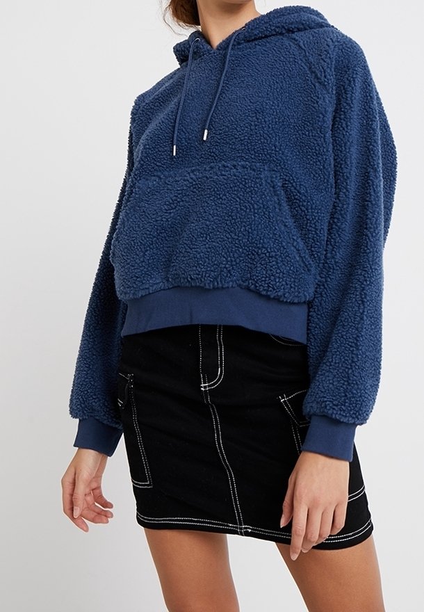 Sudadera corta de forro polar azul con capucha, cordón y bolsillo frontal. Combinada con una falda mini de mezclilla negra con costuras blancas.