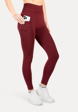 Leggings de talle alto en color burdeos con un bolsillo lateral. Están confeccionados con una tela elástica, tienen una textura suave y un diseño ajustado, combinados con zapatillas blancas.