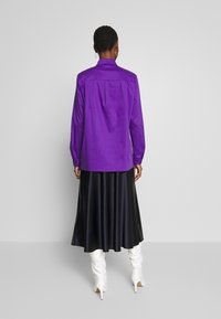 Lila långärmad skjorta med en bakfåll, i kombination med en svart kjol. Outfiten har vita högklackade stövlar och en mjuk textur.