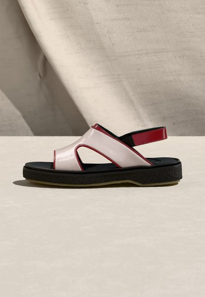Πλάγια όψη ενός λευκού σανδαλιού με κόκκινη περίμετρο, ανοιχτή μύτη, μαύρη σόλα και ρυθμιζόμενο κόκκινο λουρί στη φτέρνα, πάνω σε ανοιχτή επιφάνεια και φόντο από ύφασμα.