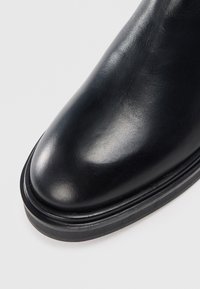 Bottine en cuir noir avec un bout arrondi, texture lisse et semelle en caoutchouc noir contrastante. Présente une couture minimale et une finition brillante.