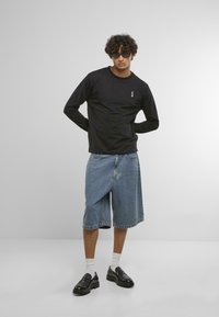 Melna garām piedurknēm krekla ar mazu logotipu, apvienots ar ziliem džinsu šortiem. Melni loaferi, baltas zeķes un tumšas brilles pabeidz apģērbu.