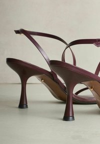 Een paar donkerviolet sandaaltjes met hoge hielen en slanke bandjes op een lichte ondergrond en een neutrale achtergrond.