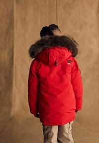Červená prešívaná parka s kožušinou podšívkou na kapucni, s prešitým logom na chrbte a nastaviteľnými popruhmi, nosená cez béžové cargo nohavice.