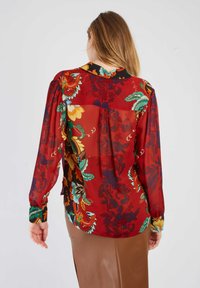 Rote Bluse mit Blumenmuster, aus transparenter Stoff gefertigt, mit langen Ärmeln und einem Kragen. Grüne und gelbe Akzente sowie eine Rückenfalte.