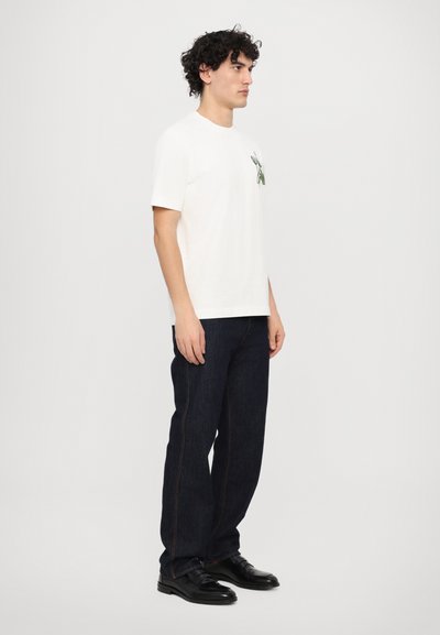 Weiße Baumwoll-T-Shirt mit kurzen Ärmeln, mit einem grünen Blumenmuster auf der Brust. Kombiniert mit dunkelblauen Jeans und schwarzen Schuhen.
