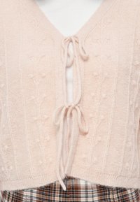 Cardigan court rose clair en maille texturée, doté d'un col en V et de fermetures à nouer sur le devant. Porté par-dessus un haut blanc et une jupe à carreaux.