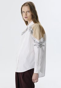 Camisa blanca con paneles beige, intrincado bordado negro en los hombros, diseño con botones, corte relajado y mangas largas.