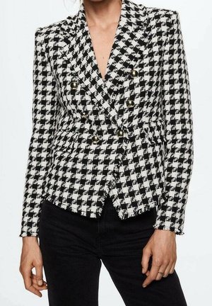 Femme portant un blazer croisé à motif pied-de-poule noir et blanc avec des boutons dorés et un pantalon noir.