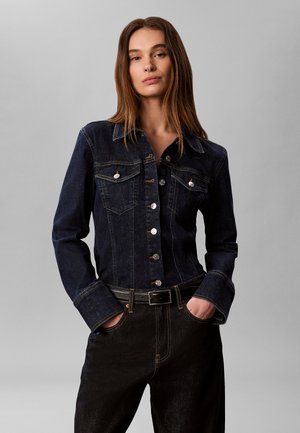 Kvinde med langt brunt hår iført en knappet mørk denimjakke og sorte bukser, står med hænderne i lommerne mod en ensfarvet grå baggrund.