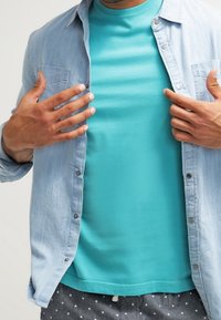 Light blue denim button-up shirt over a teal t-shirt. The t-shirt has a smooth texture, and the shirt features two chest pockets.