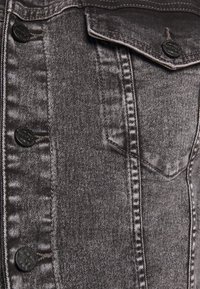 Primo piano di una giacca di jeans grigia con bottoni neri, cuciture verticali e una tasca con pattina e bottone.