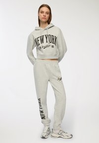 Jasnoszara, krótka bluza z kapturem z czarnym napisem "NEW YORK", w zestawie z dopasowanymi dresami i czarnymi akcentami. Miękka tekstura, luźny fason.