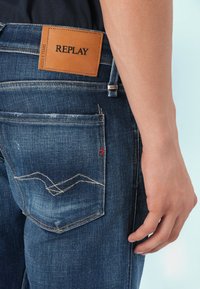 Blå denimjeans med et lædermærke mærket "REPLAY." Har en falmet tekstur, kontrasterende syninger og et lille rødt broderet logo-detail.