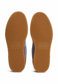 Sohlen blauer Sneakers mit braunen Gummilaufsohlen, die ein strukturiertes Diamantmuster aufweisen und das "Tommy Jeans"-Logo in der Mitte jeder Sohle tragen.