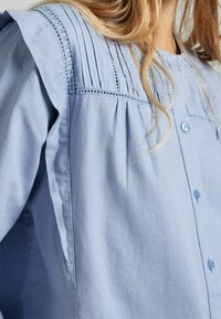 Hellblaue Bluse mit Knopfverschluss, mit Falten und Spitzenbesatz entlang der Passe. Der Stoff hat eine weiche, glatte Textur.