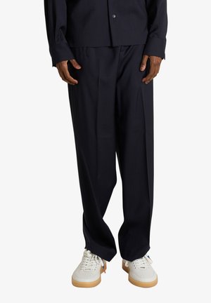 Persona che indossa pantaloni neri larghi e sneakers bianche con suola in gomma, in piedi davanti a uno sfondo bianco semplice.