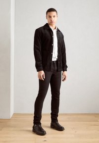 Chaqueta de pana negra con bolsillos frontales, llevada sobre una camisa blanca y pantalones negros ajustados, combinada con botas negras hasta el tobillo sobre un suelo de madera.