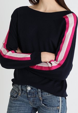 Osoba ubrana w granatowy sweter z różowymi i białymi paskami na rękawach, ze skrzyżowanymi ramionami, w połączeniu z niebieskimi dżinsami.