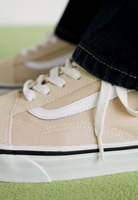 Beige lerret sneakers med hvite snøringer og en hvit side stripe. Har en teksturert såle og en svart kant langs bunnen.