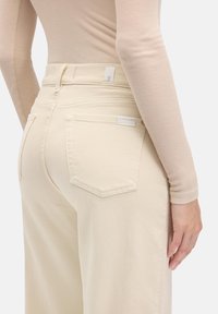 Roomkleurige jeans met een hoge taille, voorzien van twee achterzakken en subtiele stiksel details. De stof oogt zacht en gestructureerd.