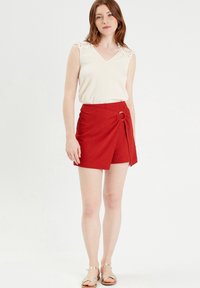 Shorts rouges enroulés avec un accent circulaire sur le côté, associés à un débardeur beige clair sans manches avec des détails en dentelle sur les épaules.