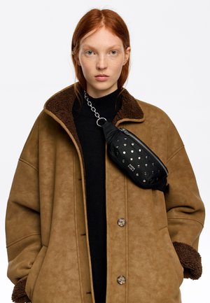 Czarna torebka na ramię ze sztucznej skóry z srebrnymi ćwiekami, zamkiem błyskawicznym i łańcuszkowym paskiem, noszona na brązowym płaszczu z kołnierzem ze sztucznego baranka.