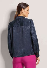 Marineblaue Bluse mit subtilm wirbelnden Muster, langen Ärmeln und Knopfdetails. Glatte Textur mit lockerer Passform, kombiniert mit rosa Hosen.