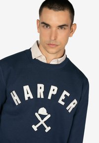 Sudadera de color navy con "HARPER" en letras bordadas blancas y gráficos de bates cruzados. Superpuesta a una camisa de cuadros con cuello.