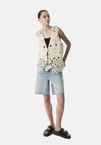 Chaleco de ganchillo en blanco roto con cortes circulares y detalles plateados, combinado con shorts de mezclilla claros y sandalias oscuras sin talón.