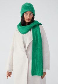 Bonnet en tricot côtelé vert et écharpe assortie en tissu doux, drapés élégamment contre un manteau de couleur claire.