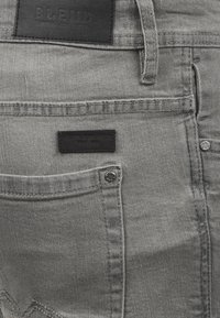 Grå denim jeans med en svart lærpatch med preget tekst. Inkluderer en frontlomme og detaljer i metall.