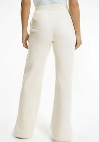 Kvinna som bär högmidjade, vida gräddfärgade byxor, står med armarna avslappnat längs sidorna mot en vit bakgrund.