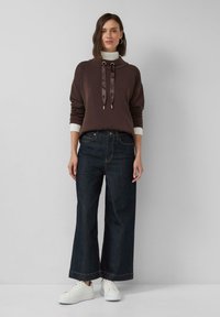 Femme debout portant un hoodie marron par-dessus un col roulé blanc, un jean large noir et des baskets blanches sur fond uni.