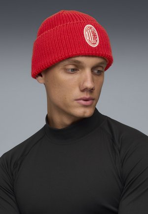 Gorro de punto rojo con textura acanalada y un logo ovalado blanco bordado. Lo lleva una modelo con una camiseta negra de manga larga.