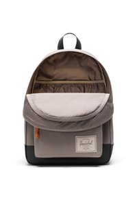 Sac à dos en tissu gris avec un dessus arrondi, une poche avant et une doublure intérieure en tan. Il comporte un écusson logo et un accent de cordon de serrage orange.