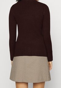 Maglione rossi a coste con maniche lunghe, indossato sopra una gonna beige a quadri. La trama è strutturata, creando un effetto visivo a strati.