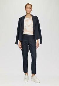 Granatowa marynarka o uporządkowanej konstrukcji, w zestawie z wzorzystą jasną bluzką i granatowymi spodniami o slim fit. Obuwie to białe trampki z grubą podeszwą.