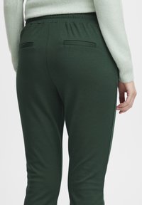 Green tapered joggers with a smooth texture, elastic waistband, back pockets, and side stripes. Paired with a light green sweater.