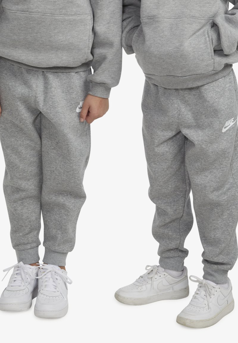 Pantaloni sport gri cu buzunare laterale și manșete elastice, combinați cu adidași albi. Prezintă logo-ul Nike alb pe pantaloni.