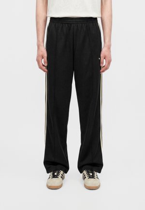 Personne portant un pantalon de survêtement noir avec des bandes blanches sur les côtés et des baskets blanches avec des lacets noirs, debout devant un fond uni.