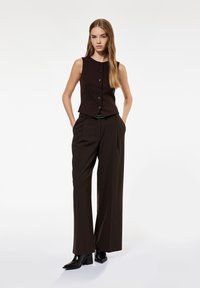 Braun ärmelloses Oberteil mit Knöpfen an der Vorderseite, kombiniert mit weiteren braunen Hosen. Schwarze Stiefeletten mit Absatz setzen einen Kontrast zum monochromen Outfit.