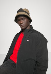 Sort letvægtsjakke med høj krave, båret over en rød skjorte. En beige og sort bucket hat fuldender outfittet med diskret branding.