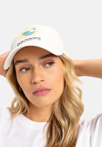 White cap featuring "BIARRITZ" logo, a colorful beach scene, and sun rays. Smooth fabric, curved brim, adjustable strap at the back.