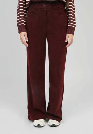 Pantalon large en velours côtelé bourgogne avec une fermeture à bouton et des poches avant, associé à des baskets blanches. Texture lisse et design ajusté.