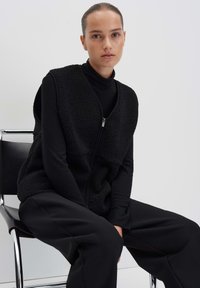 Gilet noir en tissu pelucheux, avec une fermeture éclair, un design superposé et un col haut. Porté par-dessus un col roulé noir ajusté et un pantalon large.
