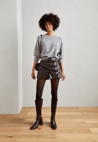 Pull en tricot gris, jupe portefeuille en similicuir noir avec des accents de boutons, collants noirs et bottes à talons bloc marron jusqu'aux genoux. Fond en parquet.