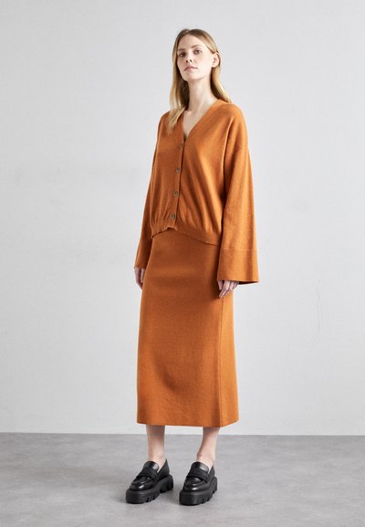 Cardigan en laine orange avec des boutons et jupe assortie. Texture lisse, design en V et manches larges. Des mocassins noirs épais complètent l'ensemble.