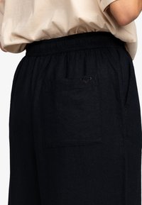 Widok z tyłu osoby noszącej czarne spodnie elastyczne w pasie z prostokątną tylną kieszenią i małym czarnym logo powyżej.